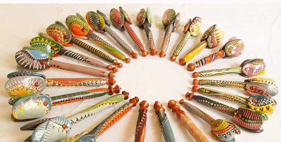 Around thirty hand-painted maracás arranged in a ring on white cloth, handles pointing inward, every gourd patterned differently.